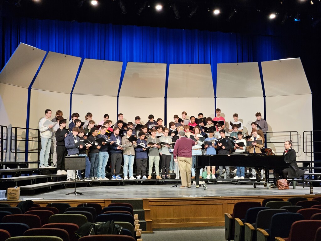 A group of male choir students is rehearsing on a stage with a conductor leading them. The students are dressed casually, holding sheet music while singing. The conductor, wearing a maroon sweater, stands in front of them, directing the performance. A pianist sits at a grand piano to the right, accompanying the choir. The stage features a curved set of acoustic panels with a blue-lit backdrop. The auditorium has rows of empty seats in the foreground.