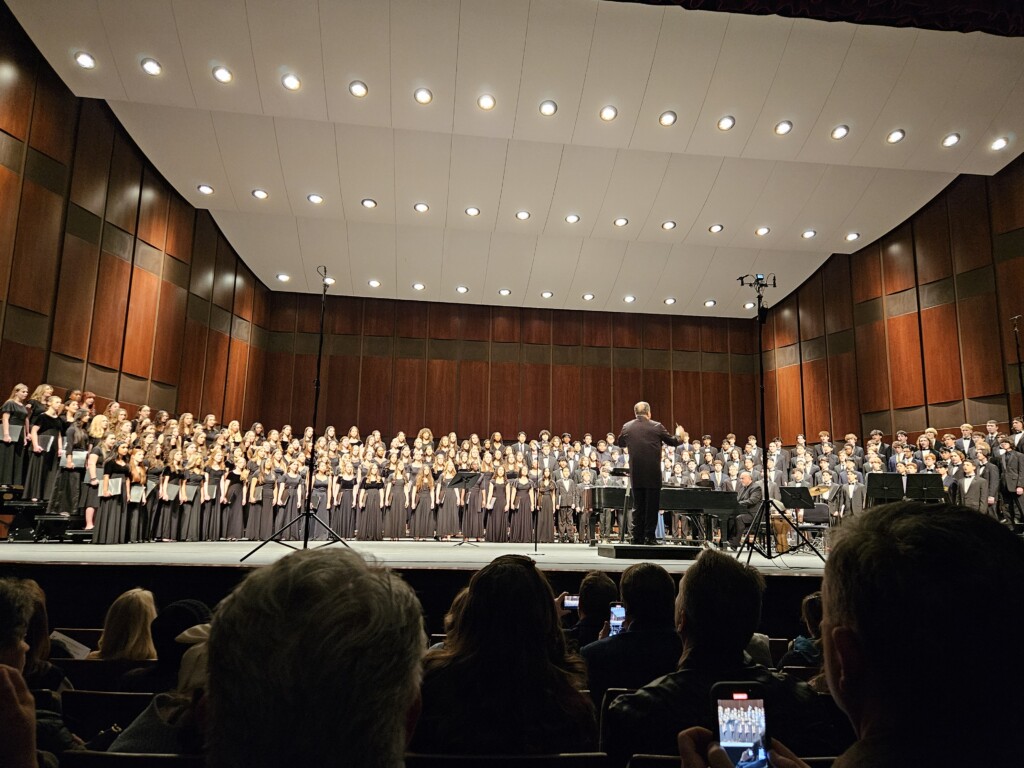 A large choir of students is performing on a well-lit concert stage with a conductor leading them. The choir is divided into two groups, with one side wearing black dresses and the other wearing black suits with bow ties. The conductor, dressed in a black suit, stands in the center, directing the performance. A pianist accompanies the choir on a grand piano. The stage has a wooden panel backdrop and a curved ceiling with recessed lighting. The audience is seated in the foreground, with some members recording the performance on their phones.